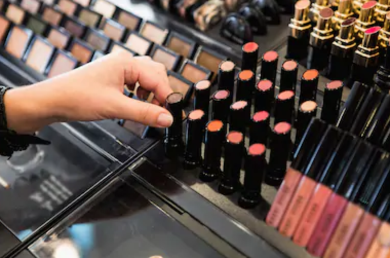 A display of lipstick and other makeup testers in a department store. A hand is reaching for a brown lipstick color.