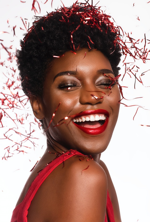 A beautiful woman with red lipstick and eye makeup is smiling while red confetti falls around her head. 