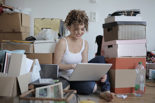 Woman surrounded by plenty of miscellaneous boxes. She is sitting on the floor barefoot and is typing on a laptop.