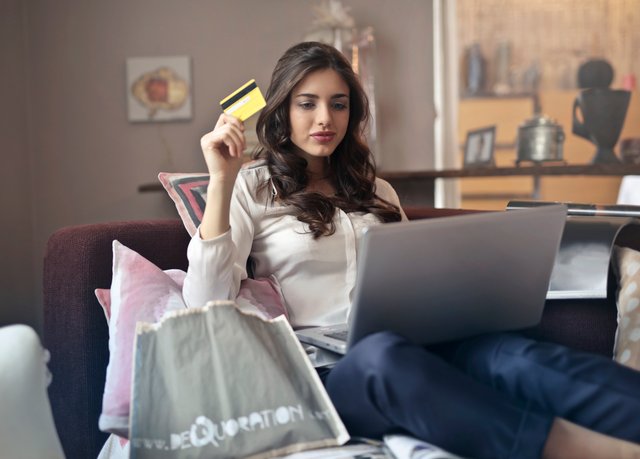 woman surrounded by shopping bags and pillows is sitting on a couch. She is looking at her laptop and holding up a debit card, implying that she is going to shop online.